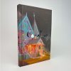 Vista frontal del libro Coraline personalizado por Skybookbindery, con la casa iluminada en tonos rosa y turquesa.